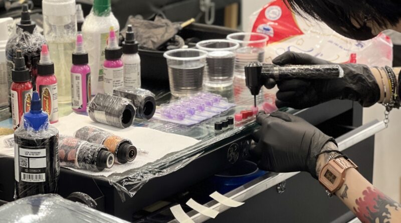 A tattoo artist in sterile gloves and mask applying a tattoo on a client's arm.